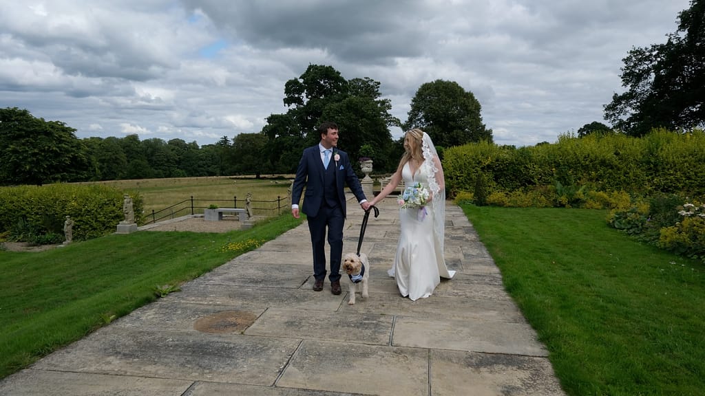 Bride and groom walking through the grounds of Lartington Hall with their dog, enjoying a relaxed moment together on their wedding day.