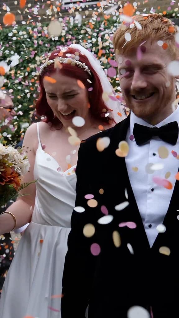 Bride and groom smiling and walking hand-in-hand through a shower of colourful confetti outside the venue, guests cheering in the background — joyful wedding celebration moment captured for Content Creation.
